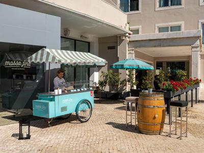 Cafetería callejera con carrito de helados azul y área de asientos frente a un hotel al aire libre soleado.