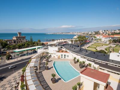 Vista sulla piscina dell'hotel, aree sedute, costa con mare e città sullo sfondo.
