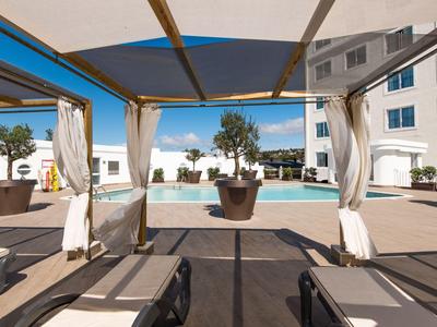 Vista attraverso un'area coperta con lettini verso la piscina dell'hotel e l'edificio in una giornata di sole.