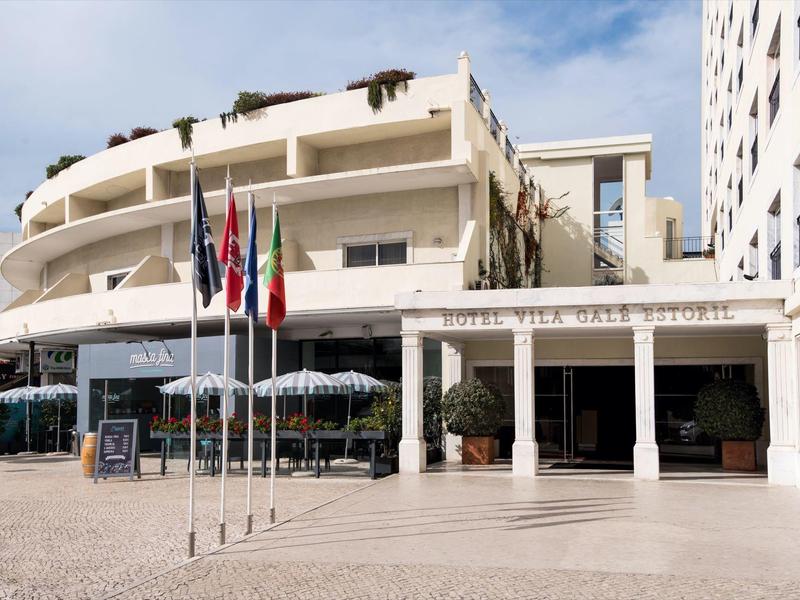 Vista exterior de un hotel con banderas, sombrillas y zona de asientos bajo cielo azul.
