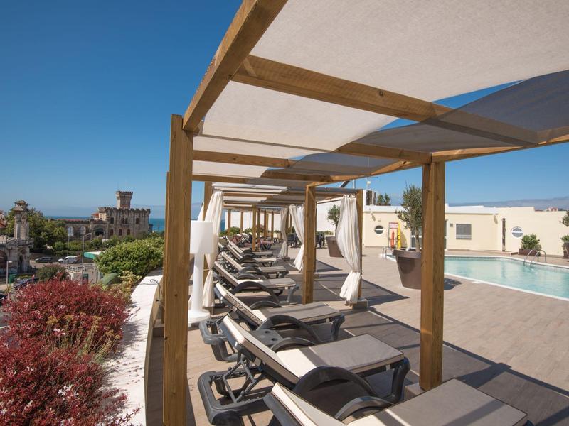 Terraza con camas de piscina y pérgola bajo un cielo despejado en un hotel de vacaciones.
