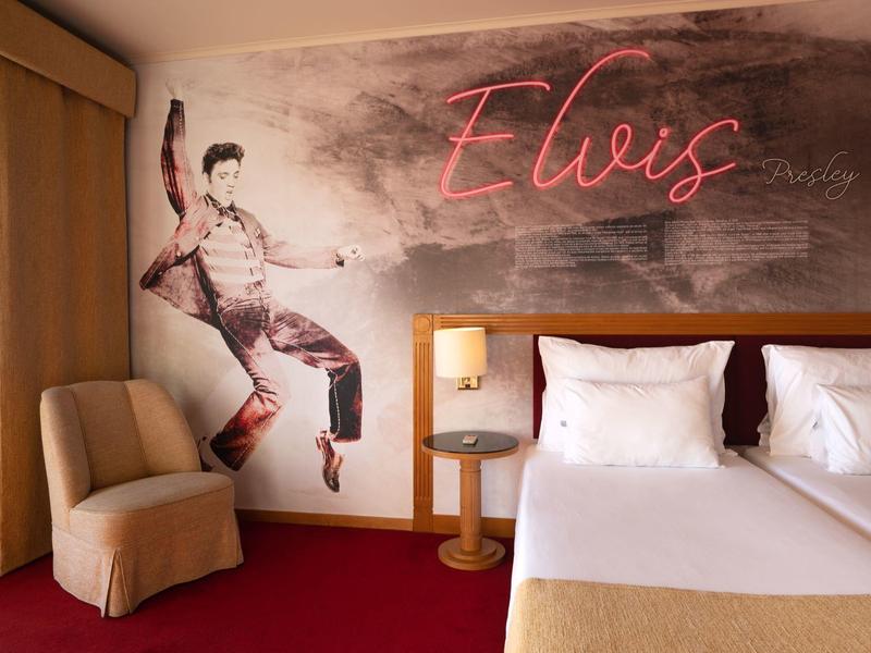 Habitación de hotel con arte mural de Elvis, cama doble, sillón beige y alfombra roja.