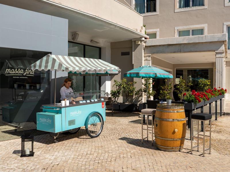 Cafetería callejera con carrito de helados azul y área de asientos frente a un hotel al aire libre soleado.