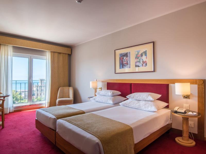 Habitación de hotel con dos camas individuales, alfombra roja y balcón con vista al mar.