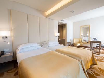 Camera d'hotel luminosa con due letti singoli, comodini e una sedia vicino a una finestra