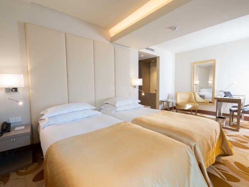 Camera d'hotel luminosa con due letti singoli, comodini e una sedia vicino a una finestra