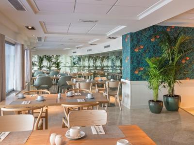 Moderner, heller Gastronomieraum mit Holztischen, Stühlen, grünen Pflanzen und großen Fenstern.