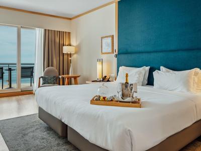 Modernes Hotelzimmer mit großem Bett, blauer Wand, Teppich, Balkon und Meerblick.