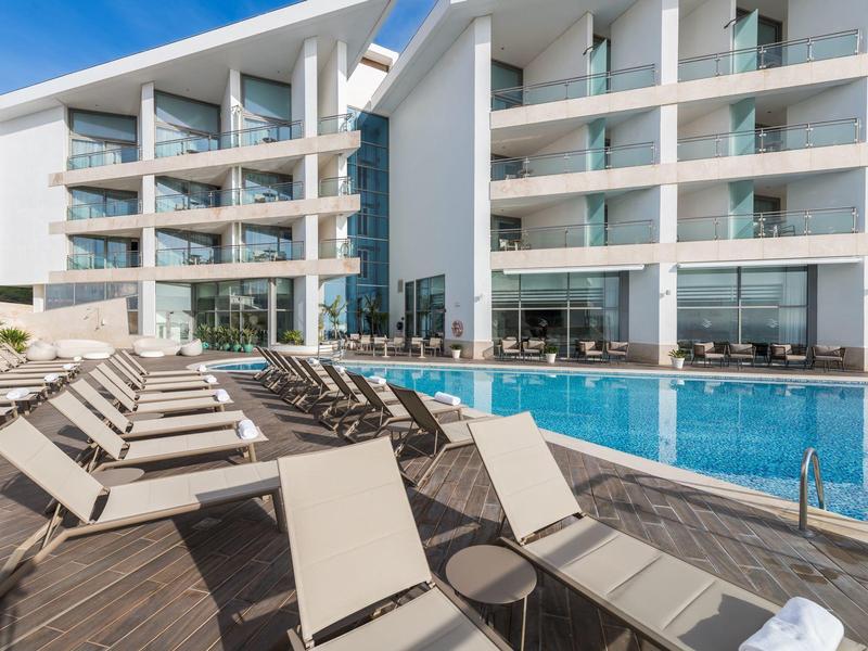 Moderner Hotelpool mit Liegestühlen auf Holzdeck und Glasgeländer vor weißem Gebäude.