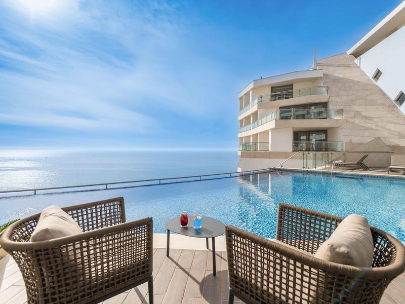 Blick auf einen Infinity-Pool mit zwei Stühlen, einem Tisch und einem weiten, blauen Himmel am Meer.