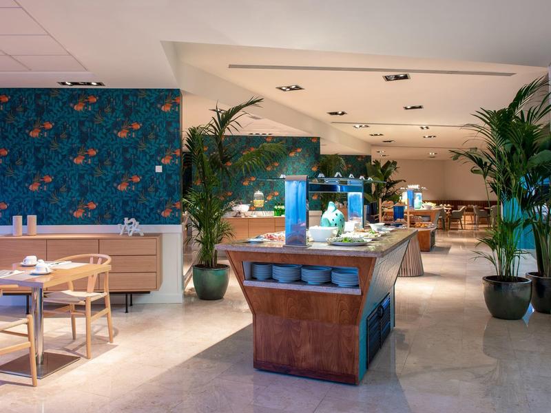 Modernes Restaurant mit hellen Holzmöbeln, großen Pflanzen und ansprechender blau-grüner Wandgestaltung.