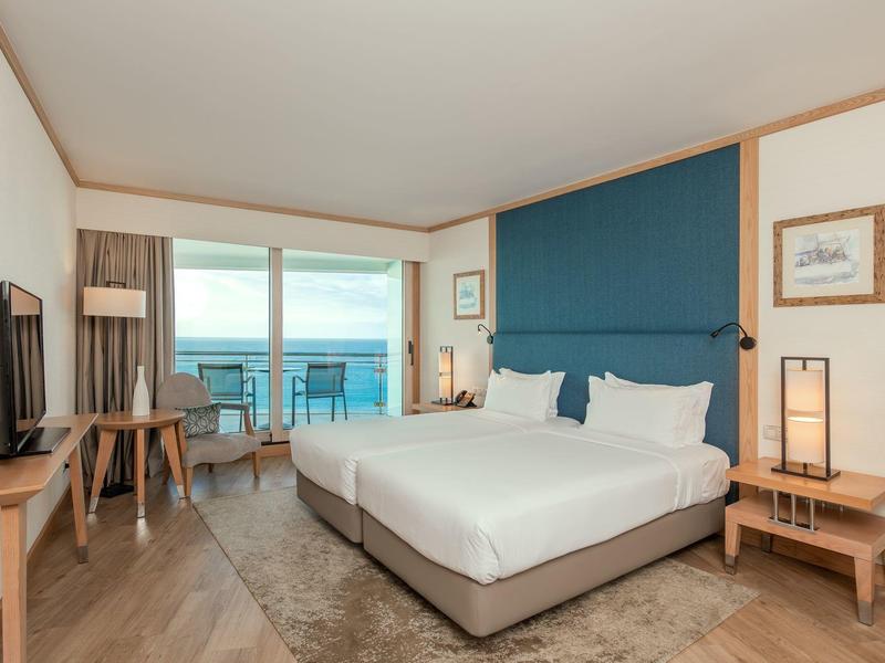 Helles Hotelzimmer mit Doppelbett, blauer Wand, Balkon und Blick aufs Meer bei Tageslicht.