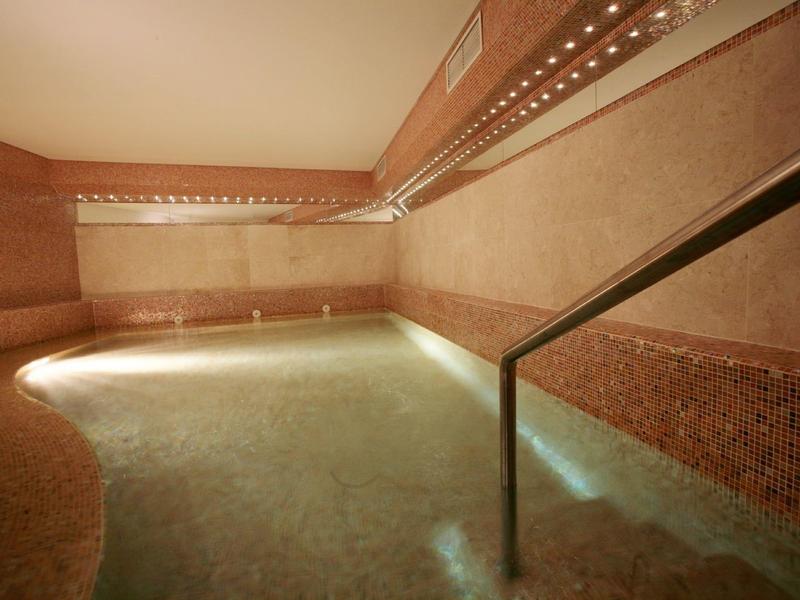 Innenraum mit rundem Pool mit warmem Licht, beige und rosa Fliesen, Edelstahlhandlauf und Wandeinbauleuchten.