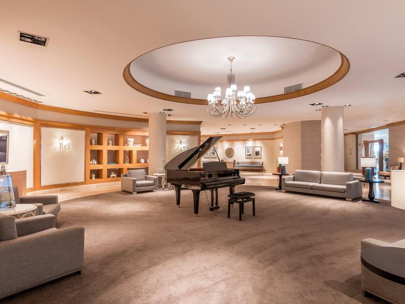 Großer Raum mit Flügelklavier, Sofa, Sesseln und Kronleuchter in warm beleuchtetem Ambiente.