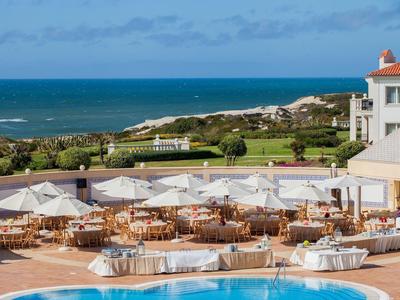 Duży taras hotelowy z stołami, białymi parasolami i widokiem na morze.