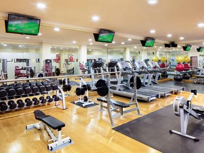 Jasna siłownia z wieloma urządzeniami, hantlami i kilkoma telewizorami na suficie.