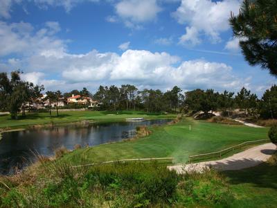 Duże pole golfowe z jeziorem, zielonymi fairwayami i budynkami w tle pod niebieskim niebem.