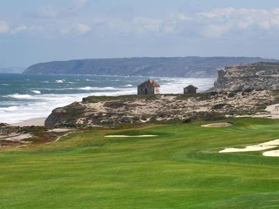 Zielone pole golfowe na skalistym wybrzeżu z morzem i chatkami w tle.