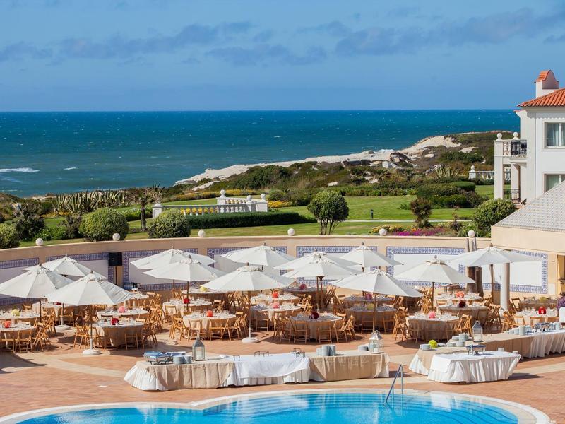 Duży taras hotelowy z stołami, białymi parasolami i widokiem na morze.