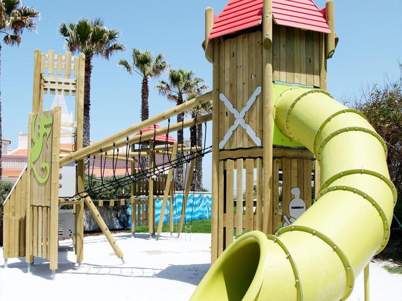 Drewniany plac zabaw z zjeżdżalnią i mostem wiszącym pod niebieskim niebem, otoczony palmami.
