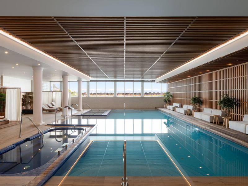 Nowoczesny kryty basen z drewnianym sufitem, leżakami i dużymi oknami z widokiem na morze.