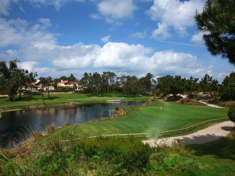 Duże pole golfowe z jeziorem, zielonymi fairwayami i budynkami w tle pod niebieskim niebem.