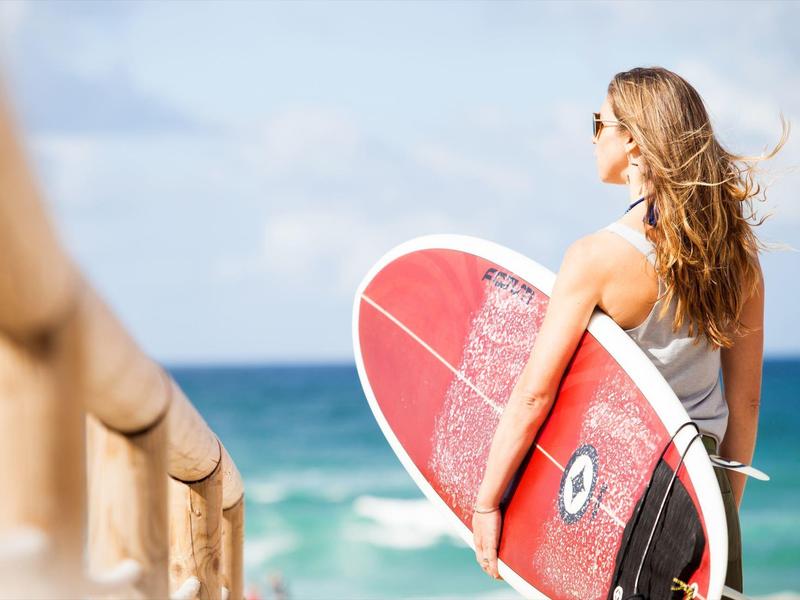 Kobieta z deską surfingową na plaży patrzy na morze pod niebieskim niebem.
