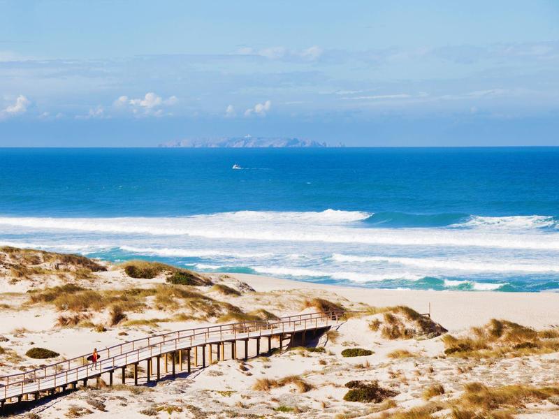 Drewniany pomost przez wydmy prowadzący na szeroką piaszczystą plażę nad błękitnym morzem.