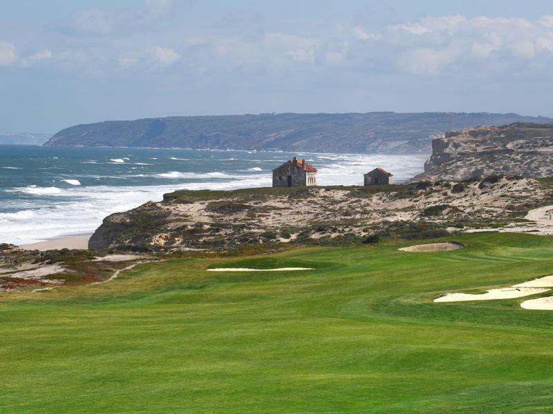 Zielony pole golfowe na skalistym wybrzeżu z falami i domami w tle.