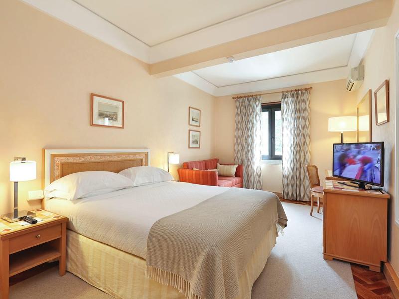 Bright hotel room with large bed, nightstands, armchair, and TV by the window.
