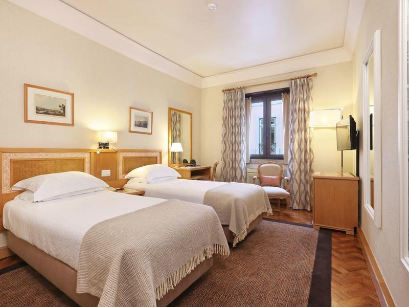 Bright hotel room with two single beds, bedside lamps, and a window with curtains.