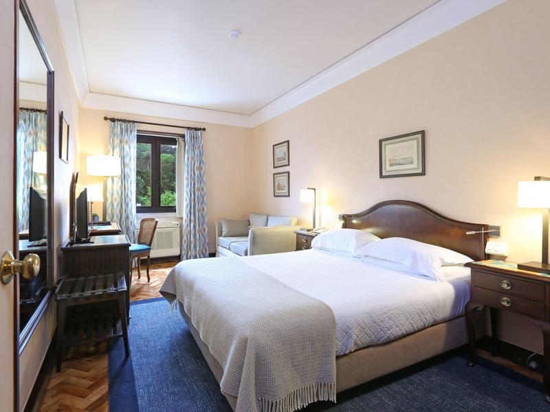 Bright hotel room with large bed, nightstands, and window overlooking greenery.