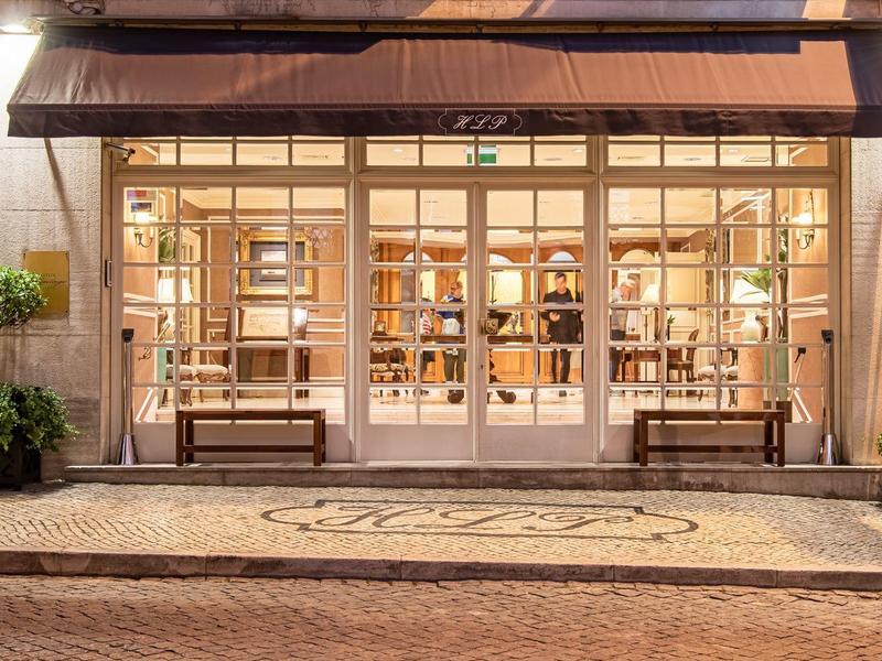 Illuminated hotel entrance with large windows, wooden benches, and plants on the facade.