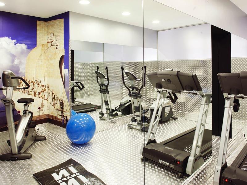 Modern fitness room with exercise equipment and large mirrored wall in hotel setting.