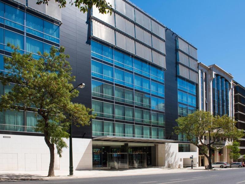 Modern hotel facade with glass front and trees along the street under clear sky