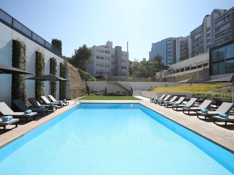 A modern outdoor pool with lounge chairs and water features, surrounded by buildings and trees.