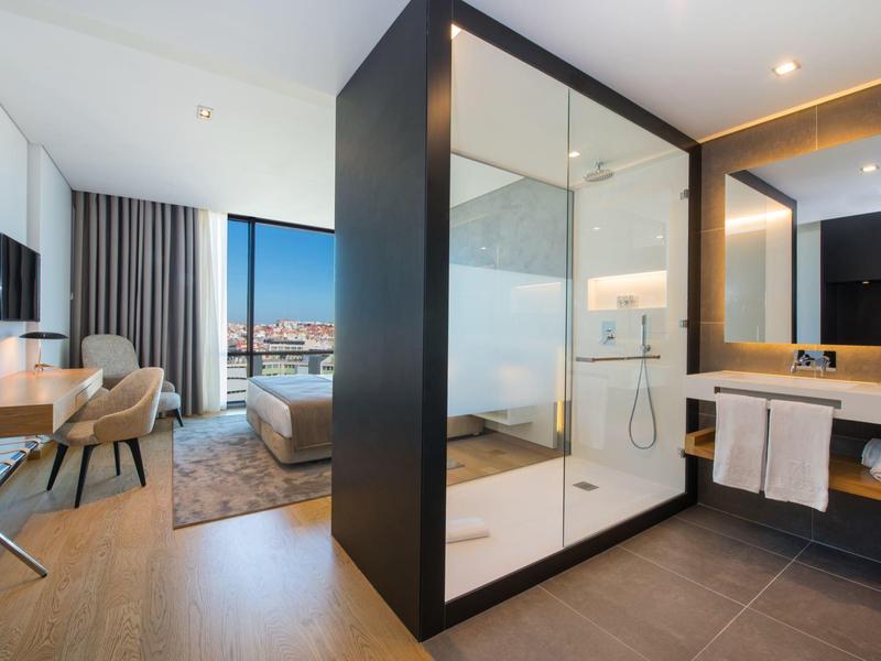 Modern hotel room with glass shower, desk, armchair, and large window overlooking the city.