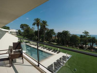 Balkon s židlemi, výhled na bazén, palmy a moře pod jasnou oblohou.