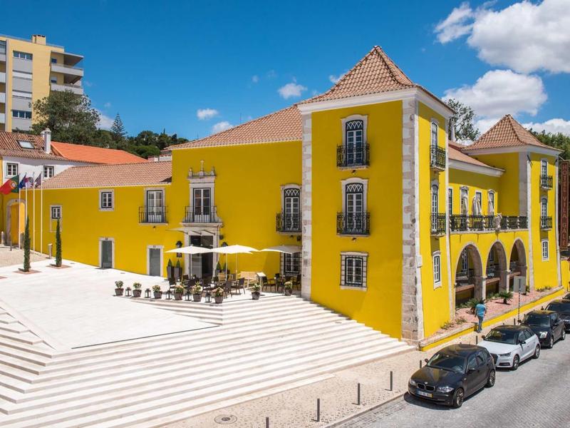 Žlutá budova hotelu s terasou a parkovištěm za slunečného dne
