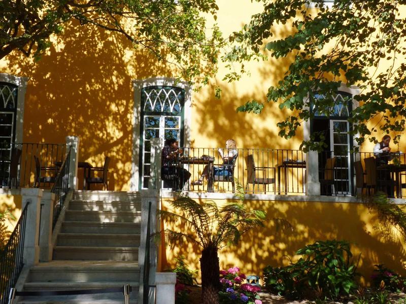 Venkovní terasa s stoly a židlemi před oranžovou budovou, obklopená stromy.