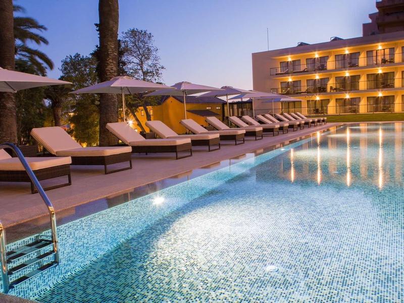 Osvětlený hotelový bazén s lehátky a slunečníky za soumraku.