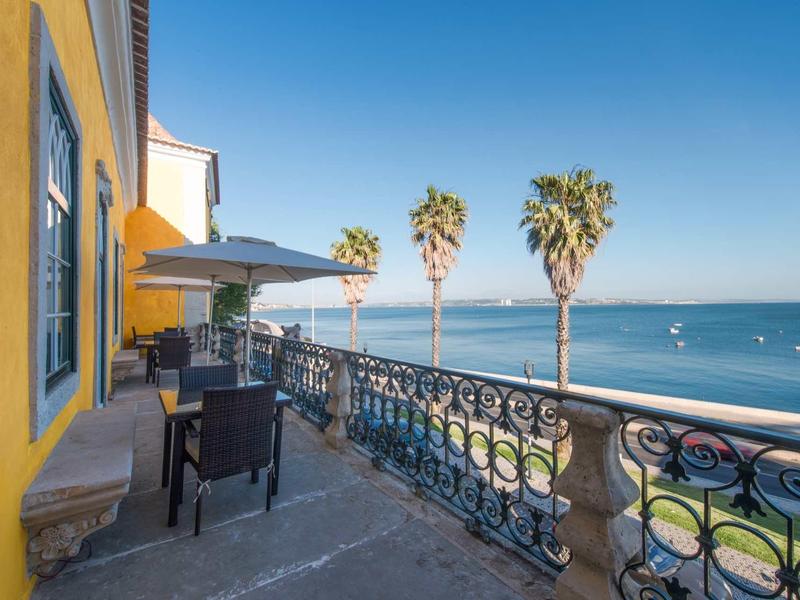 Balkon s tabulkami, židlemi a slunečníkem s výhledem na moře a palmy.