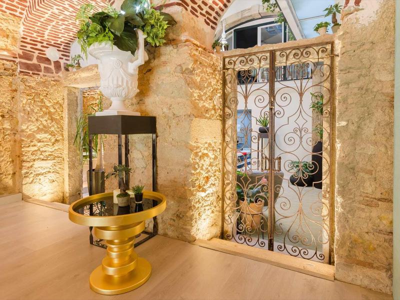 Interno luminoso con muro in pietra, cancello decorativo in metallo e tavolo dorato con piante.