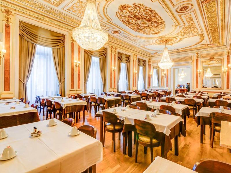 Sala da pranzo elegante con grandi finestre, lampadari e tavoli apparecchiati.