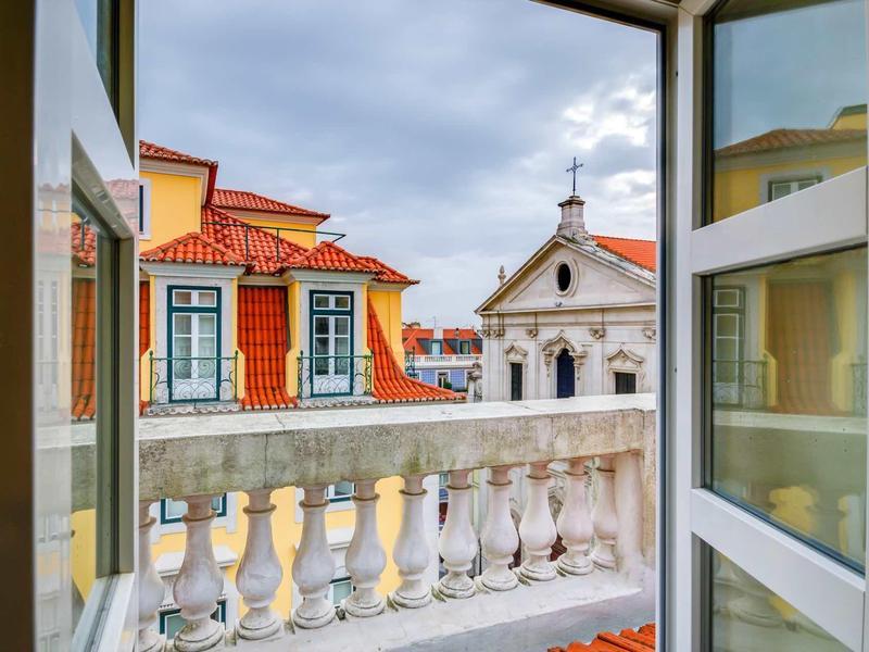Vista da una finestra aperta su un balcone con balaustra e edifici storici con tetti rossi.