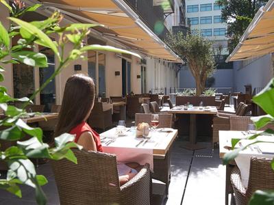 Woman sits on restaurant terrace with tables and chairs, surrounded by plants.