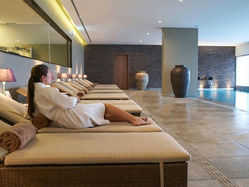 Woman in bathrobe relaxing on lounge chair beside indoor pool in modern spa area.