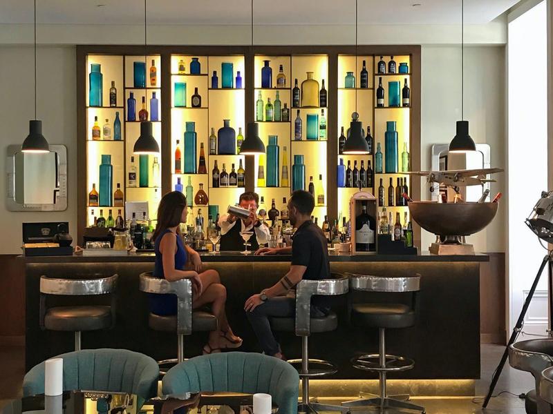 Modern hotel bar with illuminated shelves, three guests, and stylish seating.