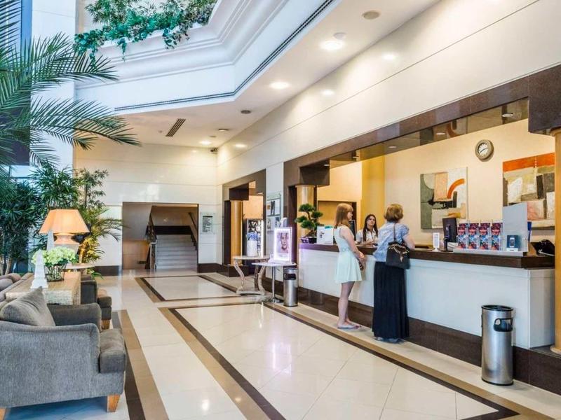 Recepción de hotel luminosa con invitados y área de descanso con sofás y plantas.