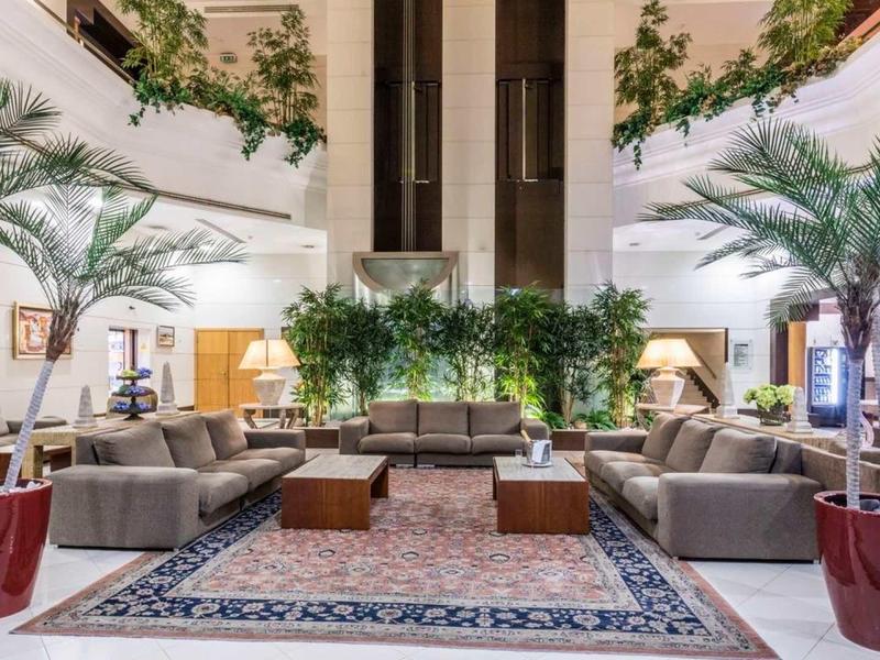 Amplio vestíbulo de hotel con áreas de descanso, plantas grandes y alfombra decorativa.